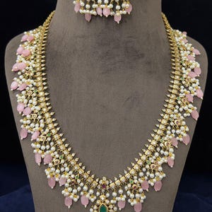 May include: A gold-toned necklace and matching earrings set. The necklace features pink teardrop-shaped beads, white pearls, and small green and red gemstones. The earrings have a similar design, with pink beads and pearls.