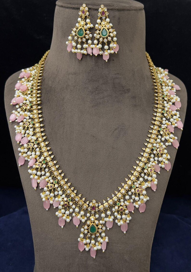 May include: A gold-toned necklace and matching earrings set. The necklace features pink teardrop-shaped beads, white pearls, and small green and red gemstones. The earrings have a similar design, with pink beads and pearls.