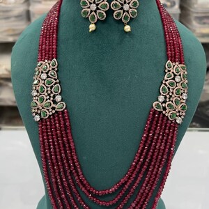 Puede incluir: Conjunto de collar y pendientes a juego. El collar presenta hilos de pequeñas cuentas rojas con cierres dorados adornados con piedras preciosas verdes y transparentes. Los pendientes tienen un diseño similar, con gemas verdes y claras.