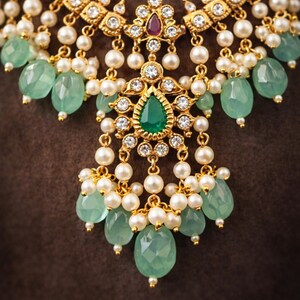 May include: Close-up of a gold-toned necklace with a central pendant featuring a large, pear-shaped green gemstone. The necklace is adorned with white pearls and smaller green gemstones, set against a brown background.