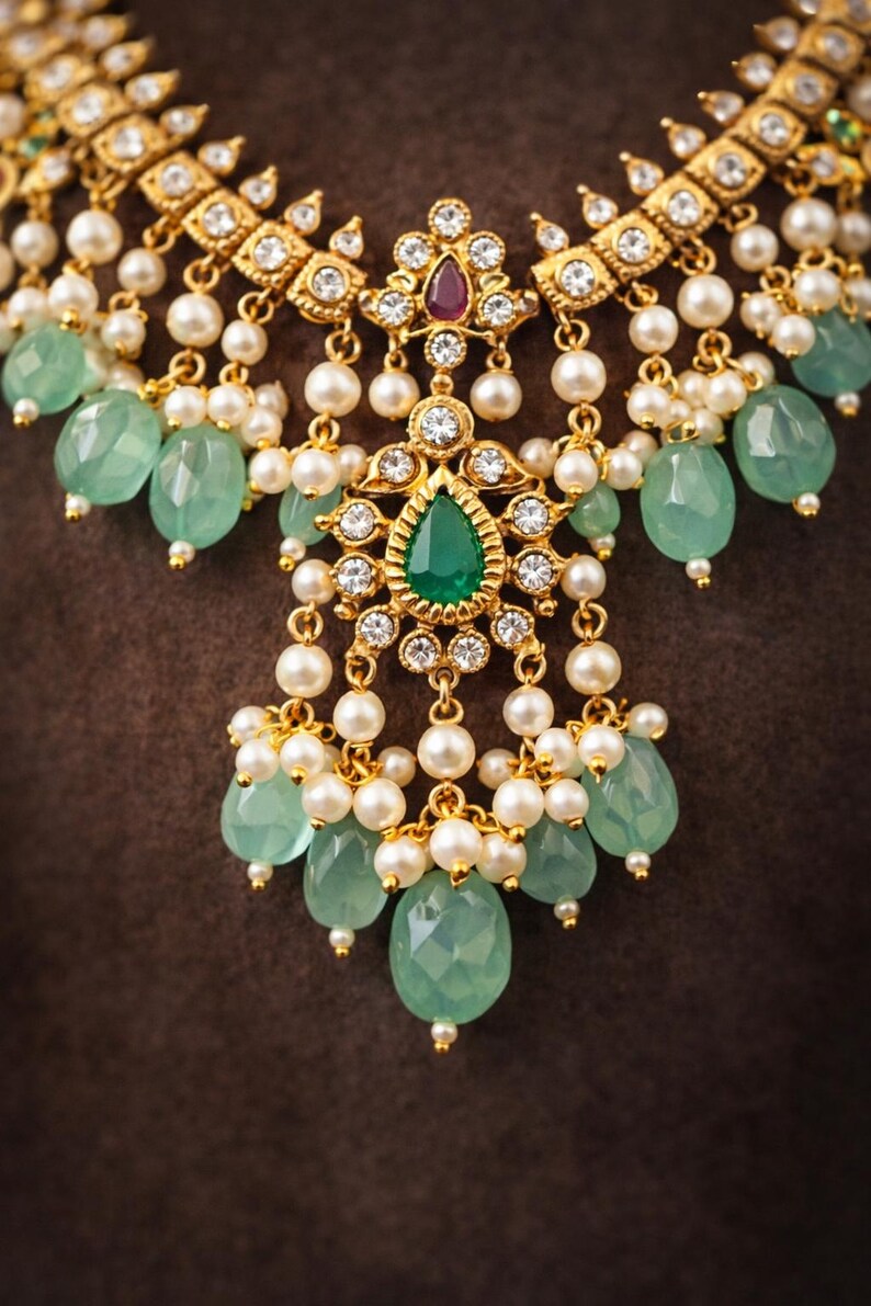 May include: Close-up of a gold-toned necklace with a central pendant featuring a large, pear-shaped green gemstone. The necklace is adorned with white pearls and smaller green gemstones, set against a brown background.