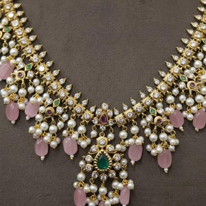 May include: A gold-toned necklace with a detailed design. The necklace features white pearls, pink gemstones, and clear crystals. It has a central pendant with a green gemstone and is set against a brown background.
