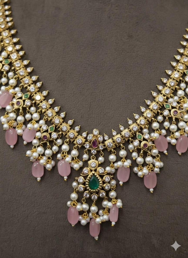 May include: A gold-toned necklace with a detailed design. The necklace features white pearls, pink gemstones, and clear crystals. It has a central pendant with a green gemstone and is set against a brown background.