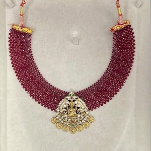 May include: A multi-strand necklace featuring deep red faceted beads arranged in a wide, curved band. At the center hangs a gold-toned pendant with intricate filigree, embedded white stones, and small circular gold coins along the bottom edge.