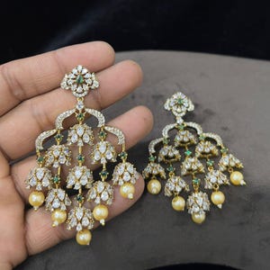 Puede incluir: Un par de pendientes tipo candelabro en tono dorado con múltiples colgantes en forma de campana incrustados con piedras blancas transparentes, pequeños acentos verdes y perlas color crema. El diseño incluye un poste floral.