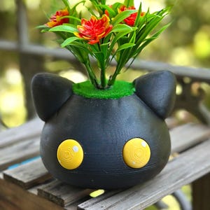 以下が含まれることがあります： 黄色い目のアクセントが付いた黒い猫型のプランターで、緑の葉を持つ赤と黄色の花が活けられています。 プランターは木製の表面に置かれ、気まぐれで装飾的なディスプレイを作り出しています。