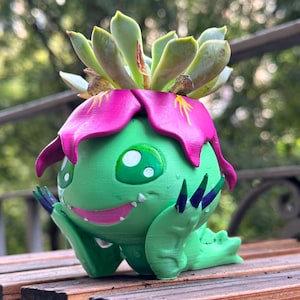 May include: A green, cartoon-like planter with a pink flower-like top and a succulent plant. The planter has large green eyes, a smiling mouth, and blue claws. It is resting on a wooden surface.