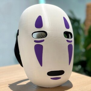 以下が含まれることがあります： 紫色のアクセントが付いた白いマスク。楕円形の目の穴と黒い口が特徴です。マスクには黒いストラップが付いており、コスプレや衣装に適しています。アニメ映画のキャラクターを彷彿とさせるデザインです。