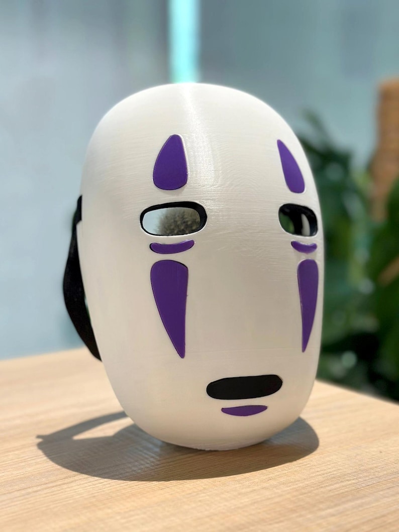 以下が含まれることがあります： 紫色のアクセントが付いた白いマスク。楕円形の目の穴と黒い口が特徴です。マスクには黒いストラップが付いており、コスプレや衣装に適しています。アニメ映画のキャラクターを彷彿とさせるデザインです。