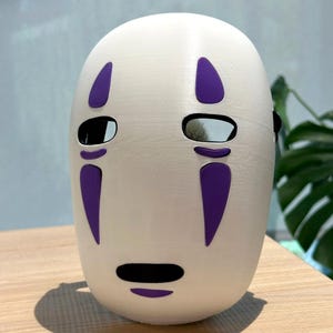 以下が含まれることがあります： 紫色のアクセントが付いた白いマスクで、目と口の切り抜きがあるミニマリストなデザインが特徴です。 マスクは滑らかで丸みを帯びた形状で、コスプレやコスチュームのアクセサリーである可能性があります。 マスクは木製の表面に置かれています。
