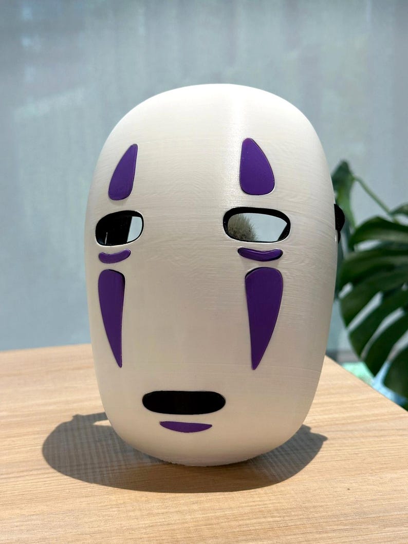 以下が含まれることがあります： 紫色のアクセントが付いた白いマスクで、目と口の切り抜きがあるミニマリストなデザインが特徴です。 マスクは滑らかで丸みを帯びた形状で、コスプレやコスチュームのアクセサリーである可能性があります。 マスクは木製の表面に置かれています。
