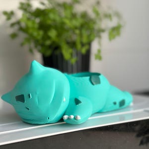 Peut inclure: Un pot de fleur Bulbasaur turquoise, représentant le personnage Pokémon avec des détails. Le pot est couché, avec sa tête et ses pattes visibles. Fabriqué en matériau lisse, il est conçu pour de petites plantes.