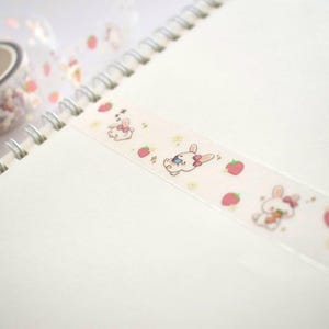 Op de afbeelding: Een rol transparante washi tape met een roze en wit ontwerp met konijnen en aardbeien. De tape is op een wit notitieboek.