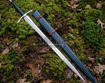 May include: A double-edged steel sword with a black leather-wrapped hilt and a crossguard, accompanied by a matching black scabbard featuring brown leather straps and metal buckles, resting on a bed of green moss and scattered brown autumn leaves.