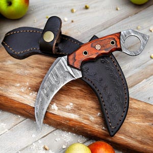Pu&ograve; includere: Un coltello karambit curvo con una lama a motivi e un manico in legno. Il coltello &egrave; in un fodero di pelle nera con un passante per cintura. L'impugnatura &egrave; di colore marrone rossiccio con tre rivetti. La lama ha un motivo ondulato unico.