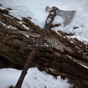 Könnte beinhalten: Eine handgefertigte Axt mit detailliertem Klingendesign und einem Holzgriff, der mit Leder umwickelt ist. Der Axtkopf weist aufwendige Muster auf, und der Griff ist mit dekorativen Elementen verziert. Die Axt liegt auf einem schneebedeckten Baumstamm.