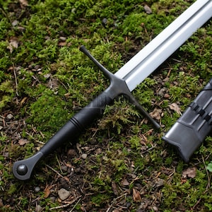 May include: A longsword with a polished silver-colored steel blade and a black textured grip. The sword features a straight crossguard and a rounded pommel with a central circular detail. A matching black scabbard lies beside the sword.