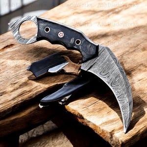 Pu&ograve; includere: Un coltello karambit curvo con una lama a motivi e un manico nero. L'impugnatura ha tre rivetti argentati e un emblema decorativo. La lama &egrave; in acciaio a motivi. Il coltello &egrave; appoggiato su una superficie di legno.