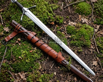 May include: A long steel sword with a silver-colored crossguard and a dark handle, positioned diagonally next to a brown leather scabbard. The scabbard features a wrap-around leather strap design with a metal buckle and a pointed metal tip.