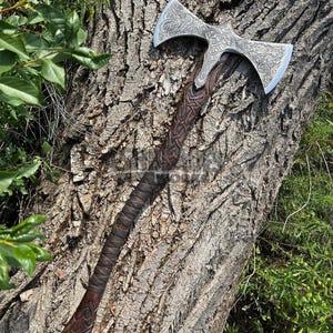 May include: A double-bladed axe with a silver head and a dark brown wooden handle. The axe head features intricate engraved patterns. The handle is wrapped in leather and has carved details. The axe is displayed against a textured tree trunk.