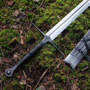 May include: A longsword with a polished steel blade, a dark matte black crossguard, and a textured black grip ending in a rounded pommel. Beside the sword lies a matching dark metal scabbard featuring black leather lacing details along its length.