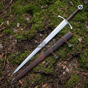May include: A silver-colored steel sword with a straight double-edged blade, a crossguard, and a dark brown leather-wrapped grip featuring a circular pommel. Beside it lies a matching dark brown leather scabbard with braided leather accents.
