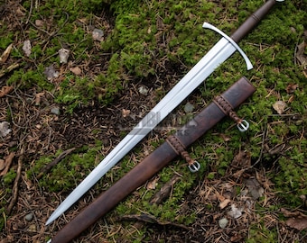 May include: A silver-colored steel sword with a straight double-edged blade, a crossguard, and a dark brown leather-wrapped grip featuring a circular pommel. Beside it lies a matching dark brown leather scabbard with braided leather accents.