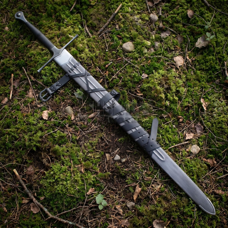 May include: A longsword with a dark metallic finish, featuring a straight crossguard and a pommel at the hilt. The blade is encased in a matching metallic scabbard, which is secured with black leather straps and a buckle for carrying.