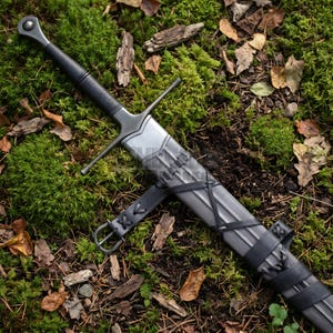 May include: A longsword with a dark metallic finish, featuring a rounded pommel, a straight crossguard, and a grip wrapped in black material. The sword is housed in a matching dark scabbard secured with black leather straps and a metal buckle.