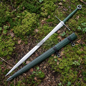 May include: A long steel sword with a double-edged blade, a silver crossguard, and a green handle featuring a circular pommel. Beside the sword lies a matching green scabbard with vertical ridges and black leather straps with metal rings.