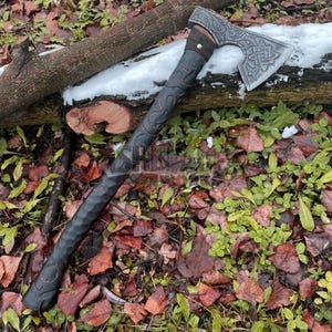 May include: Axe with a dark handle and a silver head, featuring intricate carvings. The axe is lying on a bed of leaves and greenery, with a log and a patch of snow in the background. The axe head has a detailed pattern.