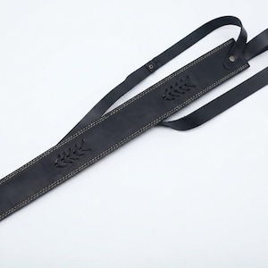 May include: A black leather sword scabbard featuring light-colored contrast stitching along the edges, decorative braided leather accents, and an integrated adjustable strap system with a metal rivet for secure attachment and carrying.