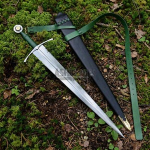 Op de afbeelding: Een stalen zwaard met een groen handvat en een pareerstang, vergezeld van een zwarte schede met een goudkleurige metalen punt. Een lange groene leren riem is aan de schede bevestigd, inclusief een metalen gesp voor aanpassing en bevestiging.