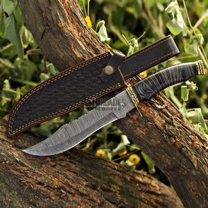 May include: A Damascus steel hunting knife with a textured blade and a dark wooden handle. The knife is paired with a black leather sheath with a woven pattern and a button closure. The knife and sheath are resting on a tree branch.
