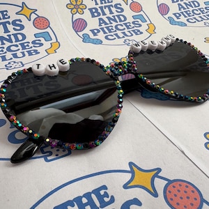 May include: Heart-shaped sunglasses with black lenses and a colourful jewelled frame. The top of the frame spells out "THE WEEKND" in white letter beads. The sunglasses rest on a paper with the text "THE BITS AND PIECES CLUB".