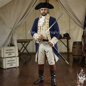 May include: A man in a historical costume, wearing a navy blue coat with gold buttons, a cream-colored vest, and tan breeches. He holds a sword. The outfit includes a black hat and tall brown boots. The background shows a tent and wooden crates.