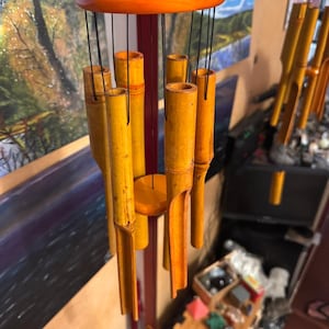 May include: A hanging wind chime with a circular orange wooden top and base. The chime features multiple bamboo tubes of varying lengths, suspended by black cords. The bamboo tubes are a warm yellow color.