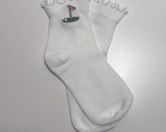 May include: A pair of white ankle socks with a ruffled top edge. Each sock features an embroidered golf green design with a flag and golf ball. The socks are made of a soft, ribbed material.