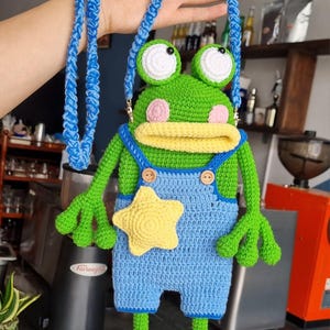 May include: A handmade crochet frog-shaped crossbody bag. The bag features a green body, blue overalls, and a yellow star. It has a blue strap and pink cheeks. The frog has large white eyes and is a whimsical accessory.