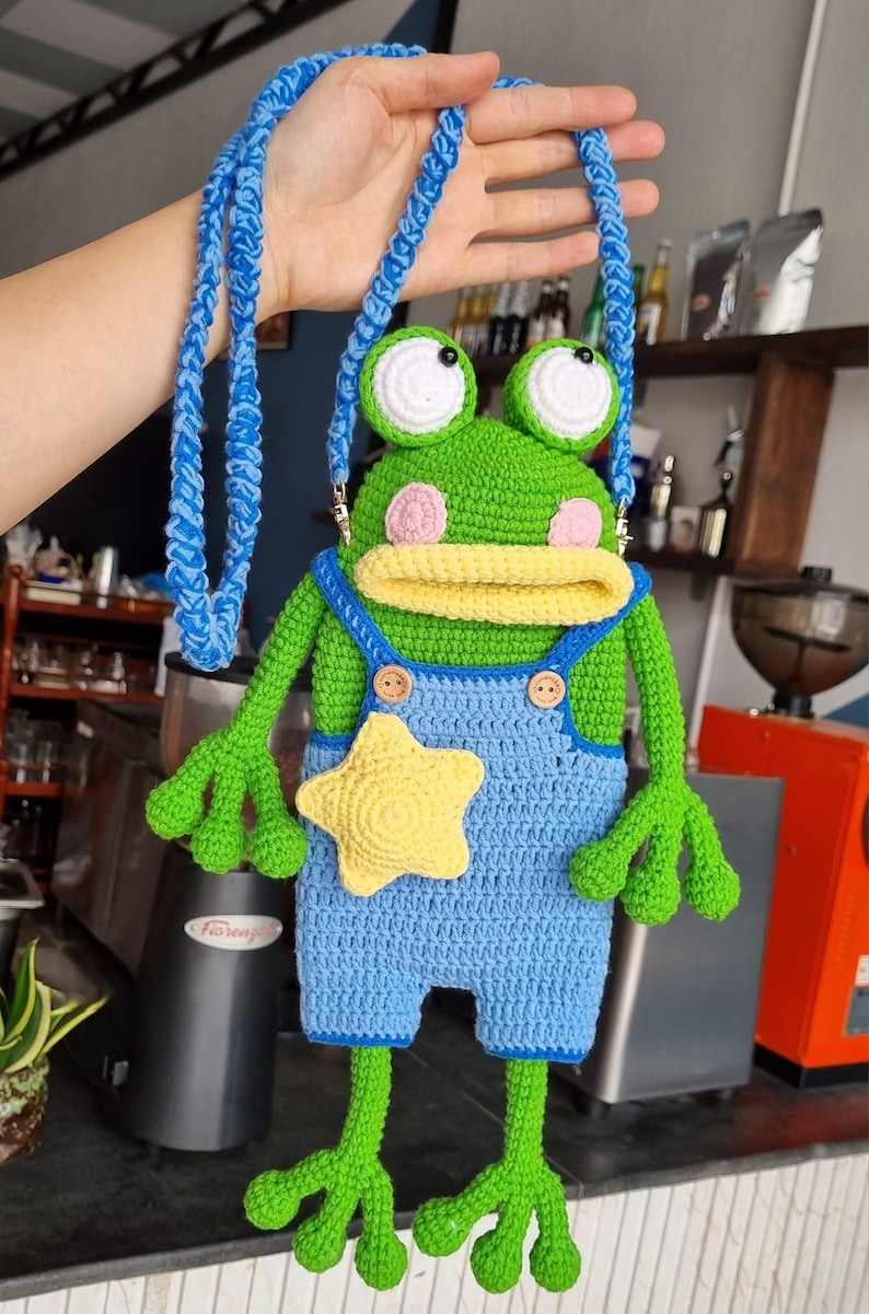May include: A handmade crochet frog-shaped crossbody bag. The bag features a green body, blue overalls, and a yellow star. It has a blue strap and pink cheeks. The frog has large white eyes and is a whimsical accessory.
