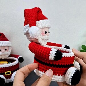May include: A handmade crochet Santa Claus figure in red, white, and black. The Santa has a red hat with a white brim and pom-pom, a white beard, and a red suit with black accents. The figure is holding a small container.