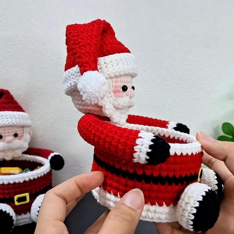May include: A handmade crochet Santa Claus figure in red, white, and black. The Santa has a red hat with a white brim and pom-pom, a white beard, and a red suit with black accents. The figure is holding a small container.