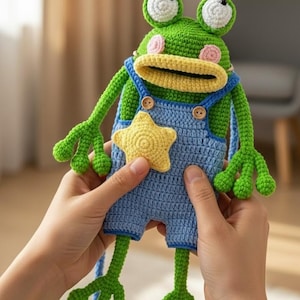 May include: A handmade green crochet frog doll with large white and green eyes, pink cheeks, and a yellow mouth. The frog wears blue overalls with a yellow star and wooden buttons. The doll is held by two hands.