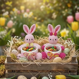 Op de afbeelding: Twee roze gehaakte konijnenfiguren met grote oren zitten in een houten kist gevuld met stro, gespikkelde eieren en snoep. De konijnen houden witte manden vast gevuld met kleurrijke verpakte snoepjes. De achtergrond is een vage tuinscène.