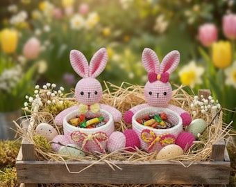 May include: Two pink crochet bunny figures with large ears sit in a wooden crate filled with straw, speckled eggs, and candy. The bunnies hold white baskets filled with colorful wrapped candies. The background is a blurred garden scene.