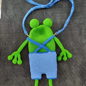 May include: A handmade crochet frog-shaped crossbody bag in green and blue. The frog has a green head with two round eyes, blue overalls, and a blue strap. The bag features arms and legs, and is designed to be worn over the shoulder.