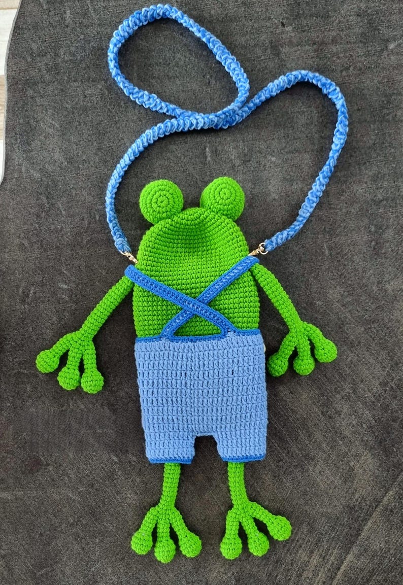 May include: A handmade crochet frog-shaped crossbody bag in green and blue. The frog has a green head with two round eyes, blue overalls, and a blue strap. The bag features arms and legs, and is designed to be worn over the shoulder.