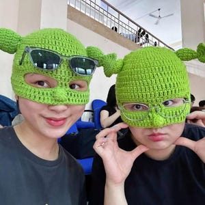 Puede incluir: Dos personas con sombreros verdes de ganchillo hechos a mano con rasgos de Shrek, incluyendo orejas y nariz. Un sombrero tiene gafas de sol, el otro gafas. Ambos llevan camisetas negras.