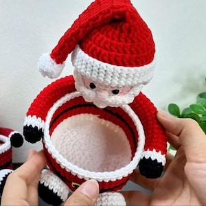 May include: A handmade crochet Santa Claus basket in red, white, black, and pink. The basket features a red hat with a white trim and pom-pom, a white beard, and a red body with black accents. The basket is designed to hold small items and is perfect for Christmas.