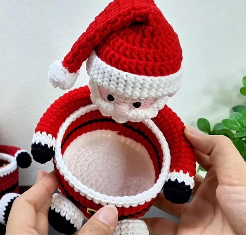 May include: A handmade crochet Santa Claus basket in red, white, black, and pink. The basket features a red hat with a white trim and pom-pom, a white beard, and a red body with black accents. The basket is designed to hold small items and is perfect for Christmas.